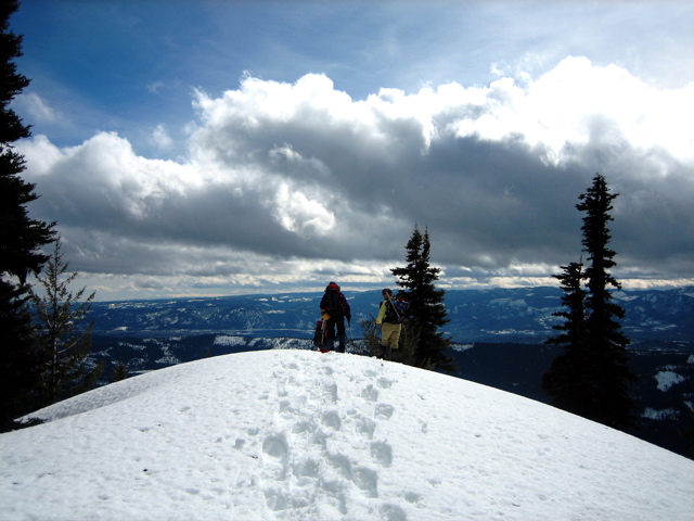 Snowshoers walk down a snowy ridge toward a puffy cloud on Yellow Hill