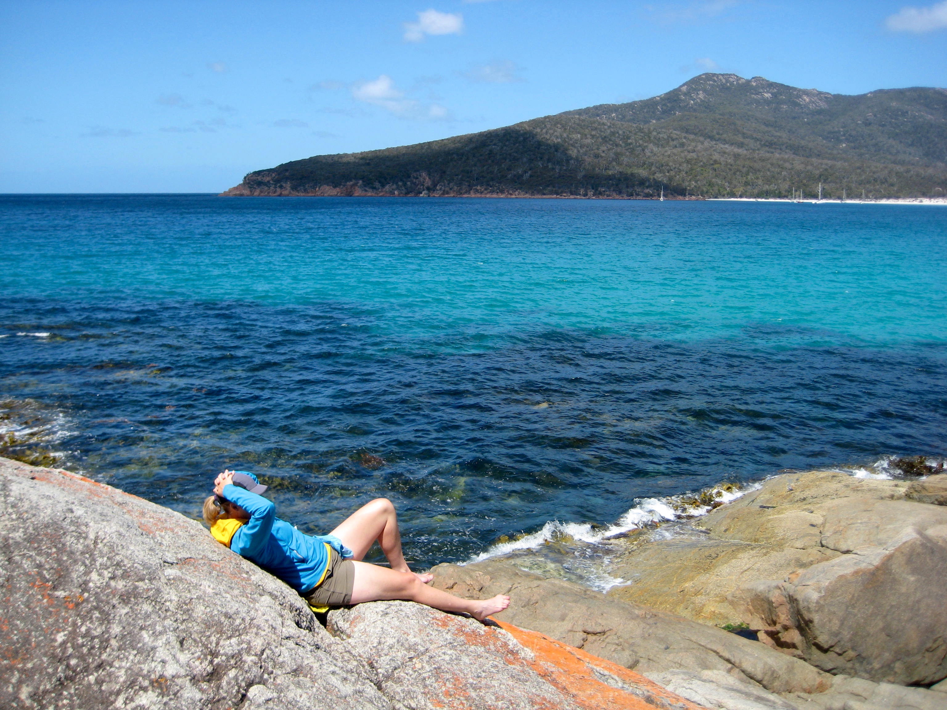 hiker laying down on the rock slabs at Wineglass Bay in Tasmania's Freycinet National Park with the blue ocean and mountain range in the background