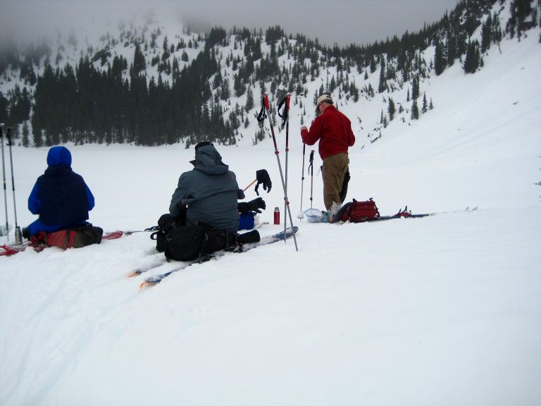 Backcountry skiers eat lunch on the shore of snowy Lake Valhalla during the Nason-Smith Ski Traverse