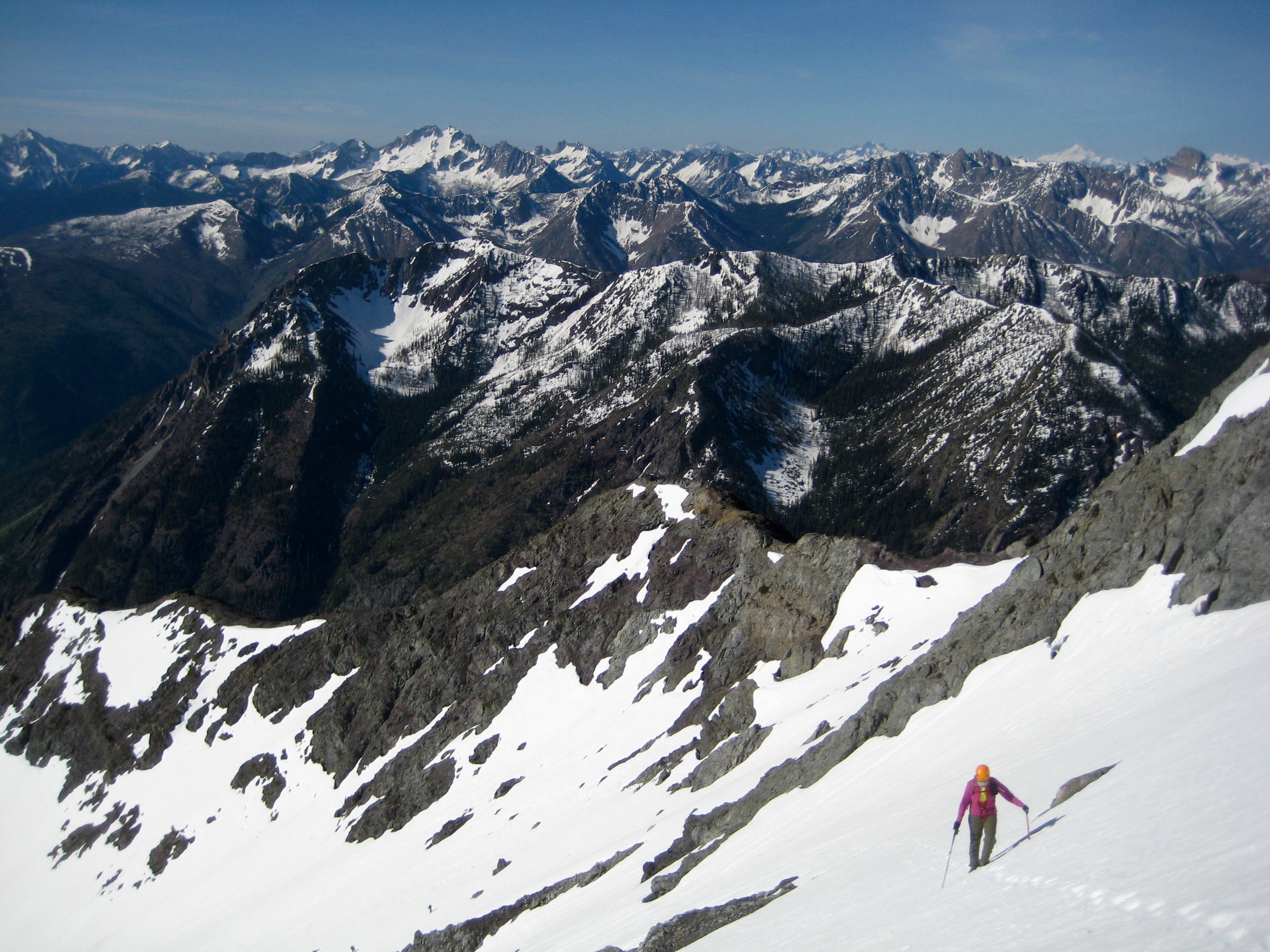 looking down on mountain climber booting up snow field with the ridge line of Robinson Mountain behind and the Pasayten Mountains in the background
