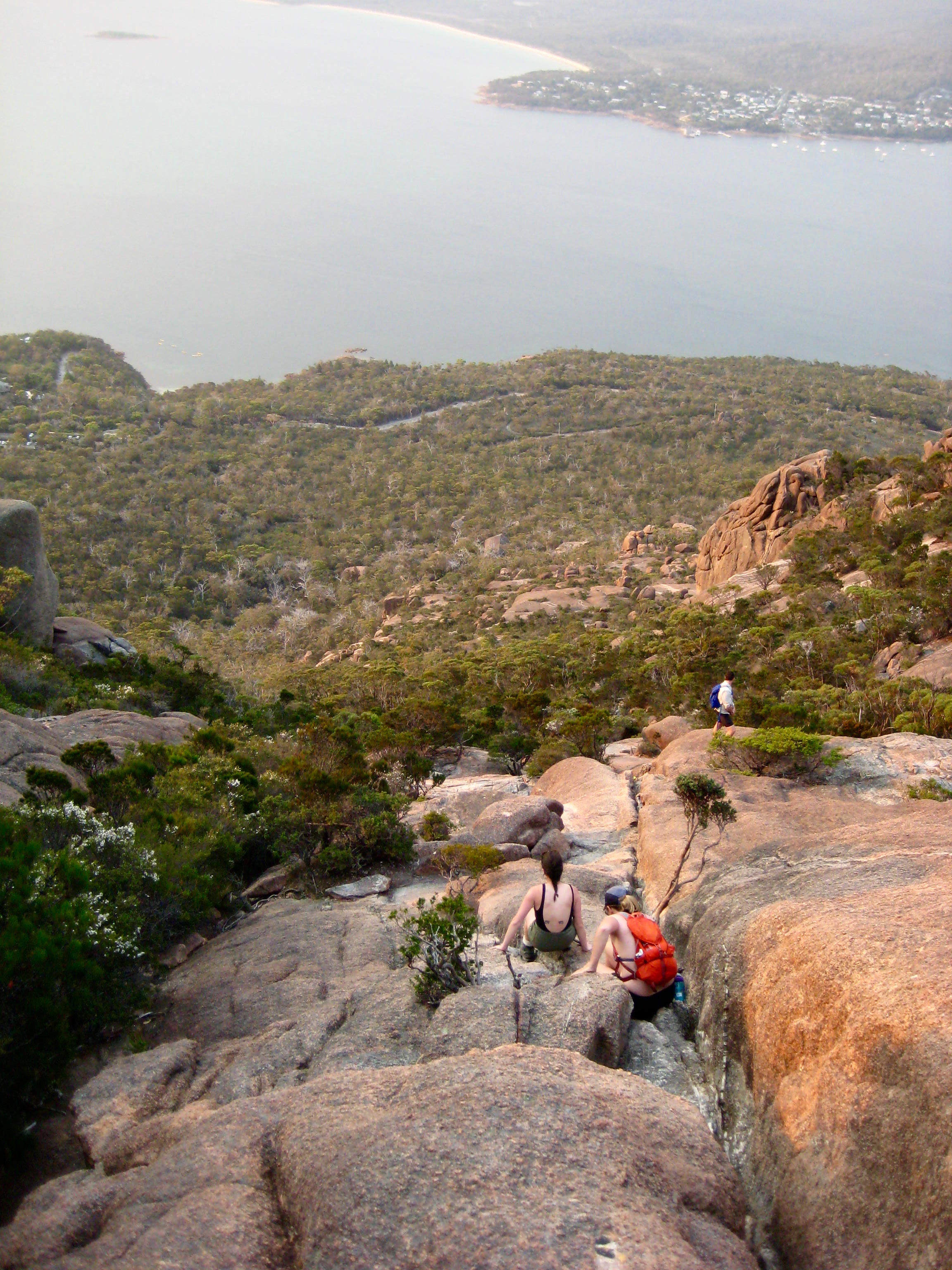 looking down on hikers carefully descending giant, rock, slab crack on Mt Amos in Tasmania's Freycinet National Park with green hillside and ocean bay below