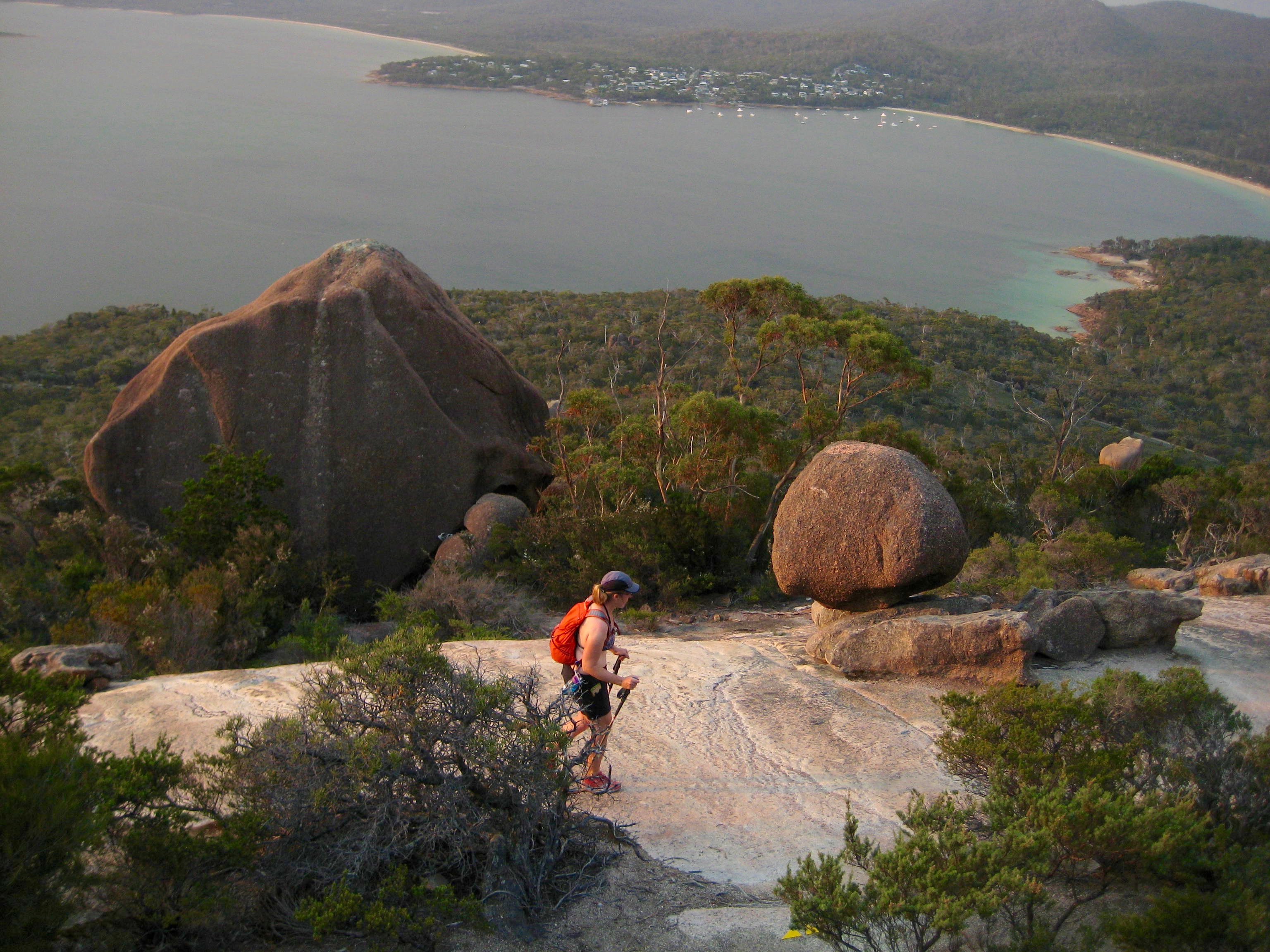 looking down on hiker descending rock slabs on Mt Amos in Tasmania's Freycinet National Park with giant boulders and ocean bay in the far distance