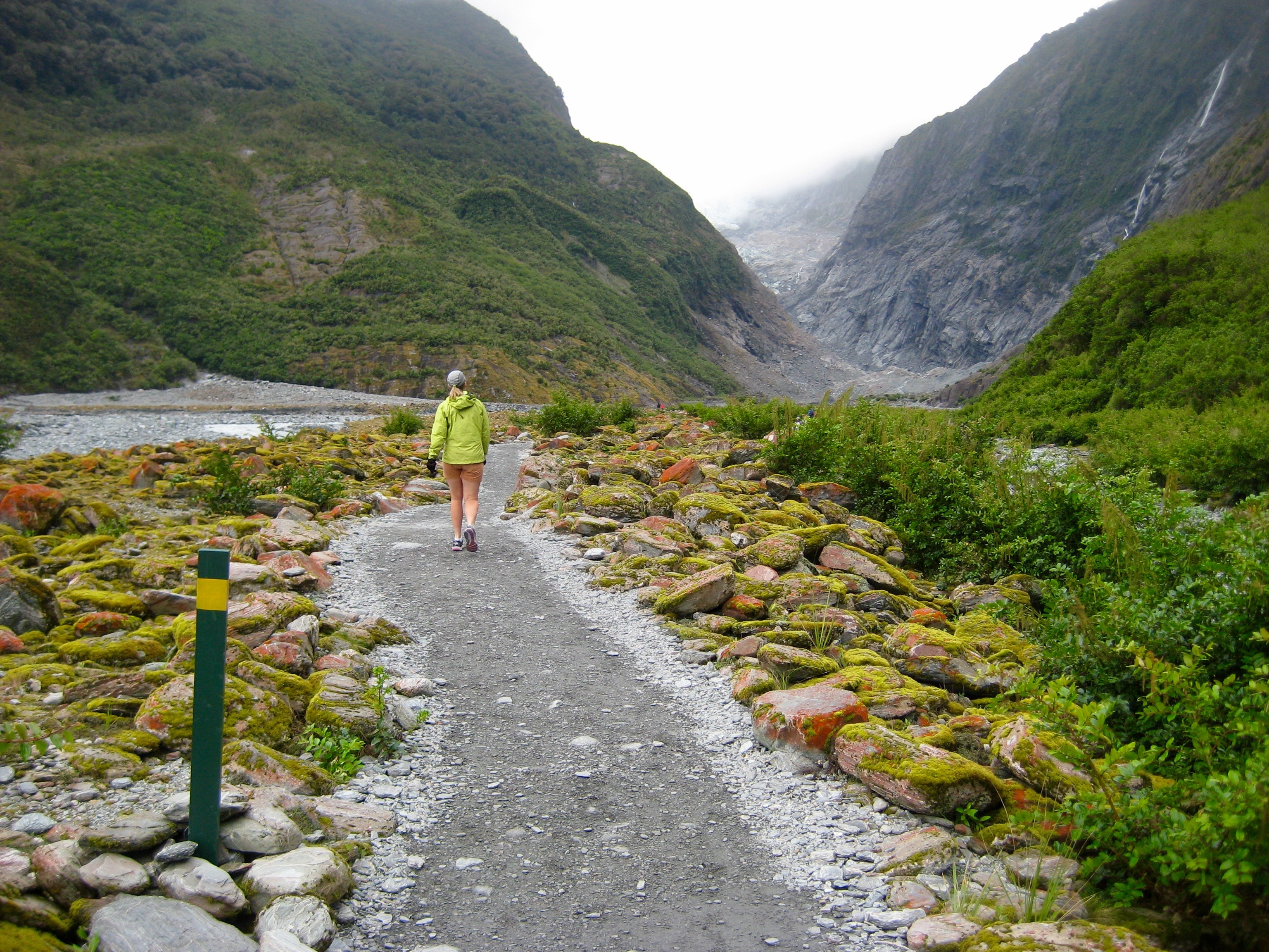 A solo hiker walks up a gravel trail in a wide glacial valley en route to Franz Josef Glacier Viewpoint