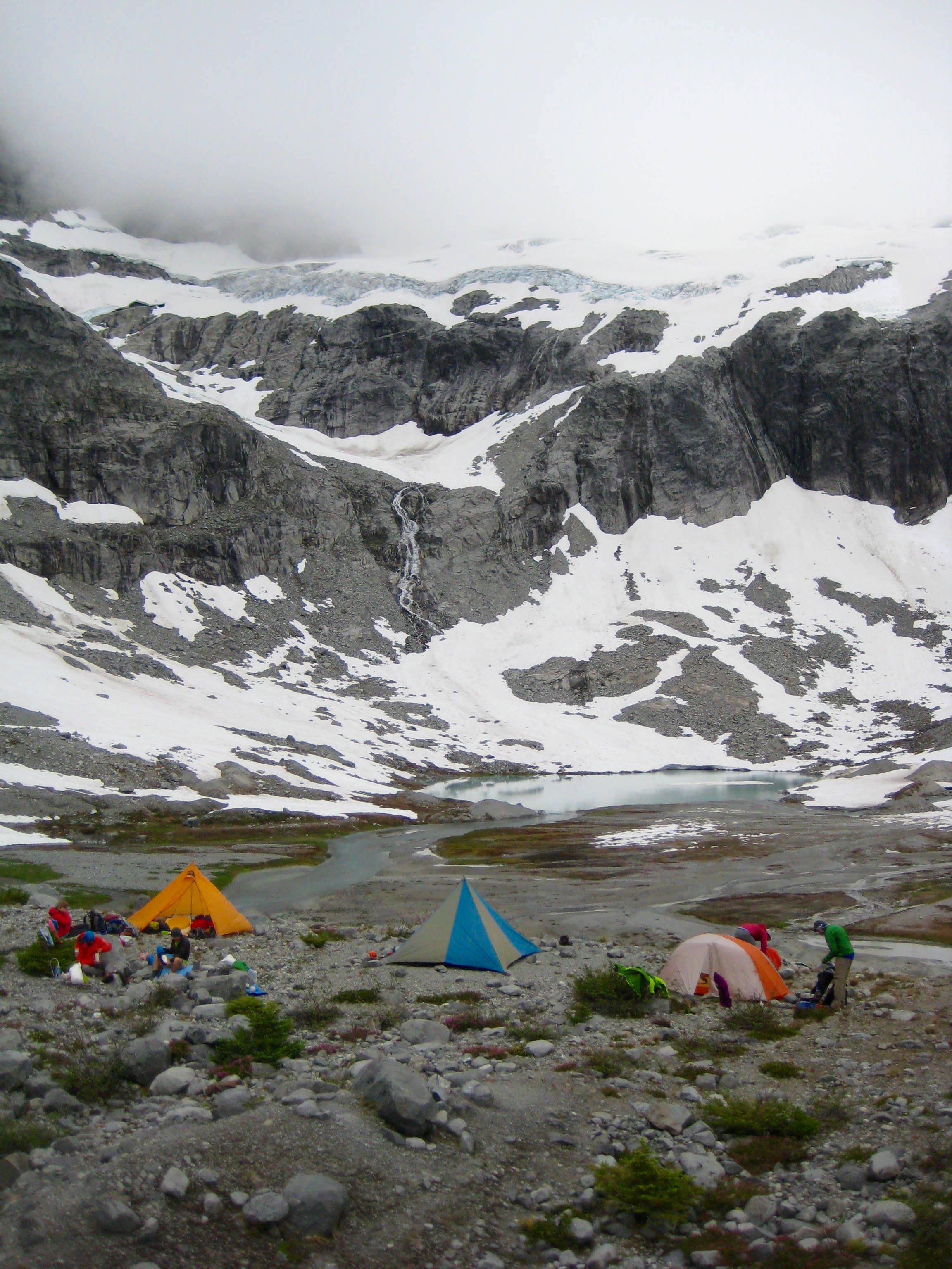mountain climber's camp at Ouzel Lake in the American Chilliwack Mountains with low cloud cover hanging over rock cliffs with linguring snow patches 