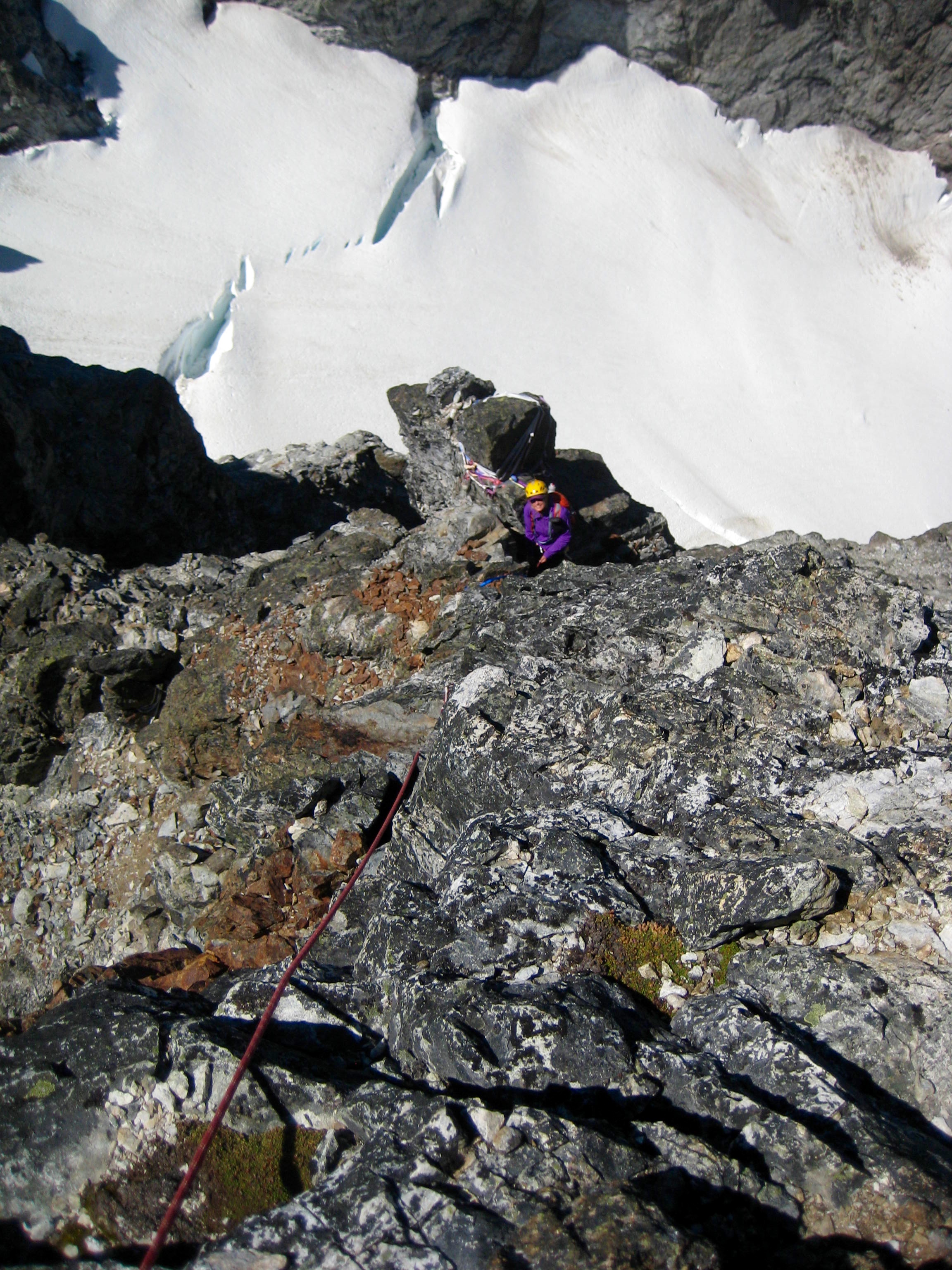 looking down at roped mountain climber belaying from rock horn on the summit tower of Hard Max Peak in the American Chilliwack Mountains with snow field below