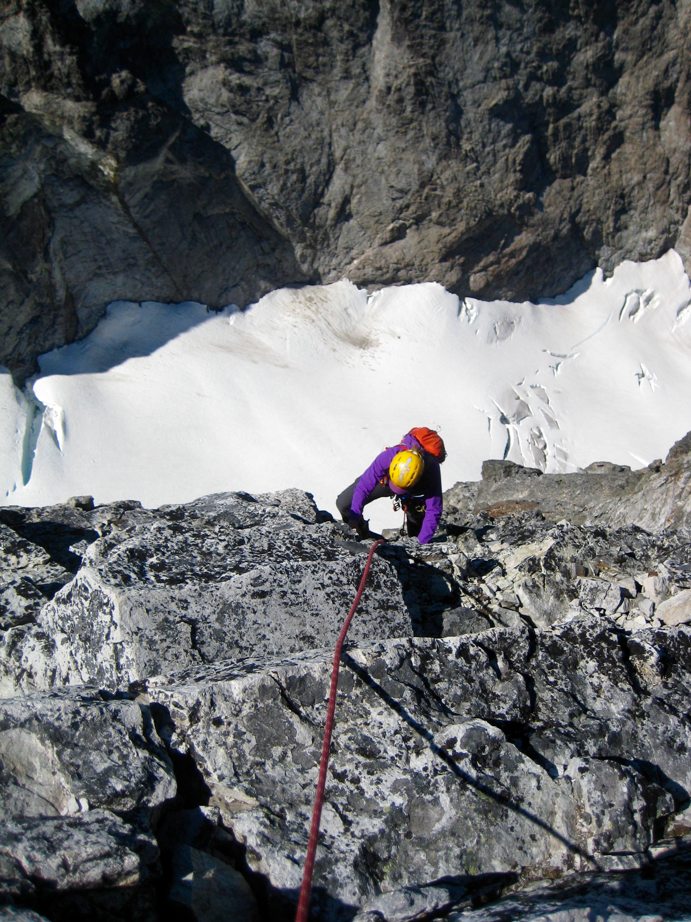 looking down on roped mountain climber scrambling the steep rock summit tower of Hard Max Peak in the American Chilliwack Mountains with snow field below