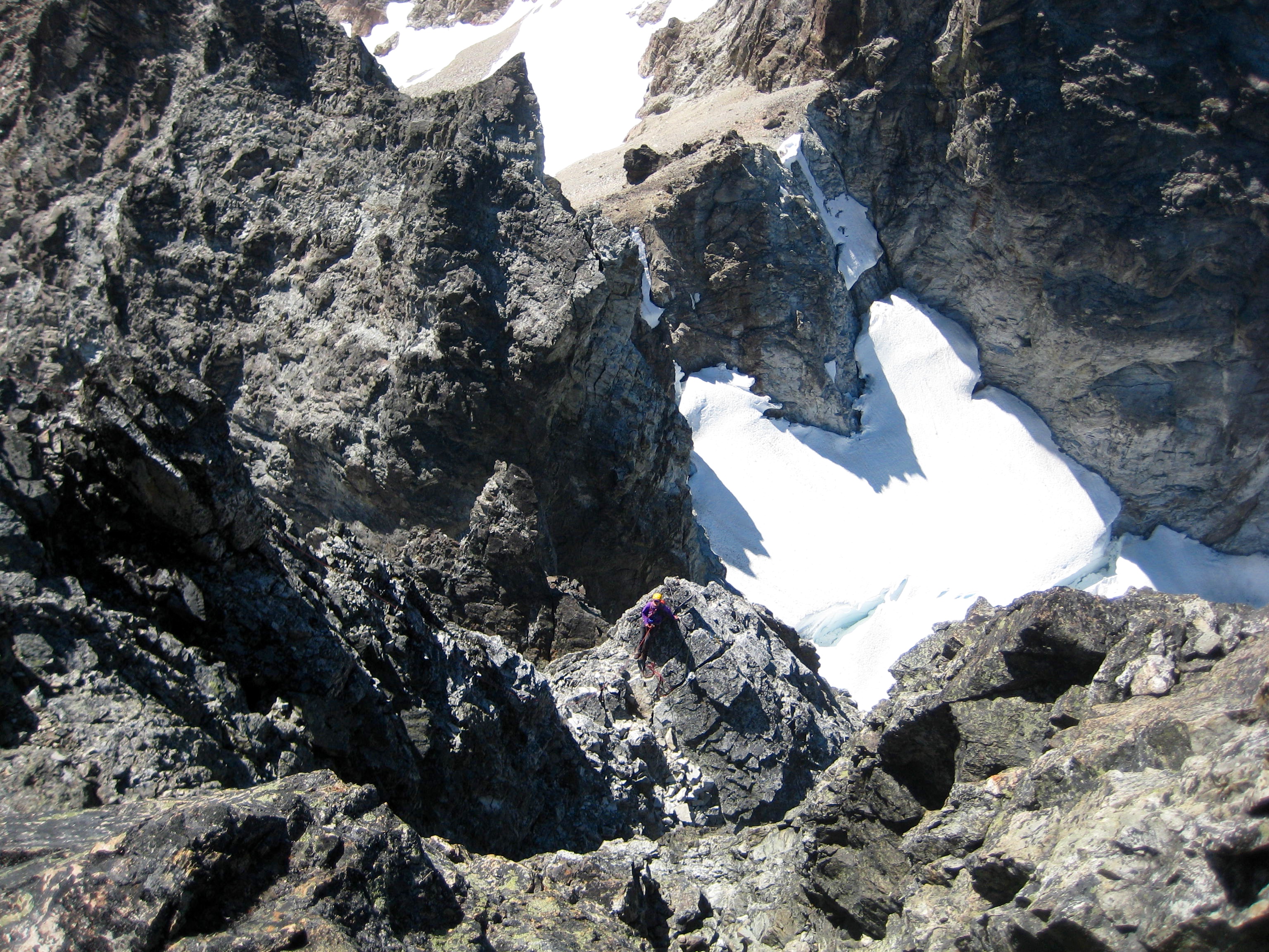 Looking down on snow field and climber belaying on third and final rock pitch of Hard Mox Peak
