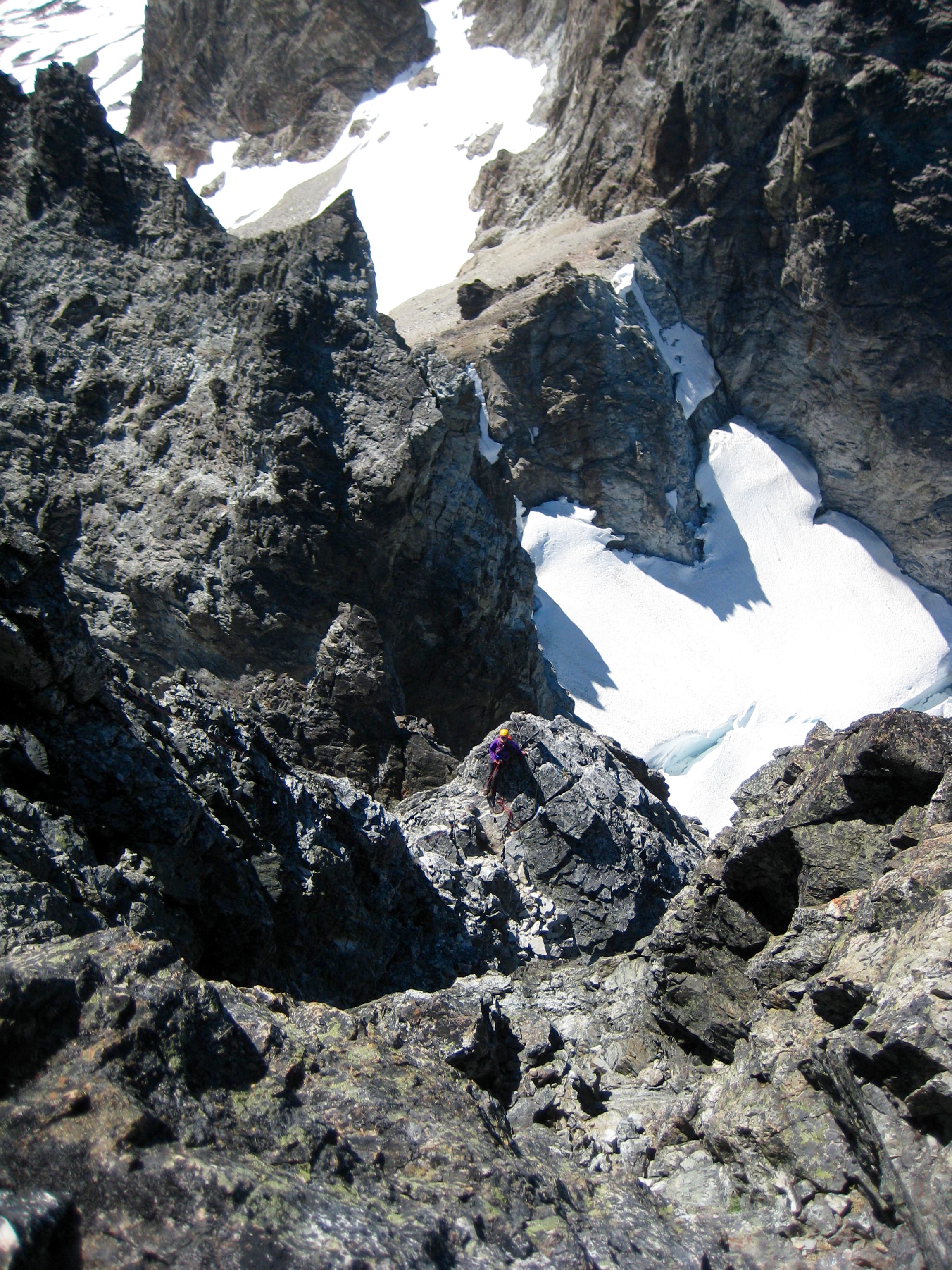 looking down on roped mountain climber in the steep rock belaying final pitch of summit tower on Hard Mox Peak in the American Chilliwack Mountains with Col Of The Wild in the background