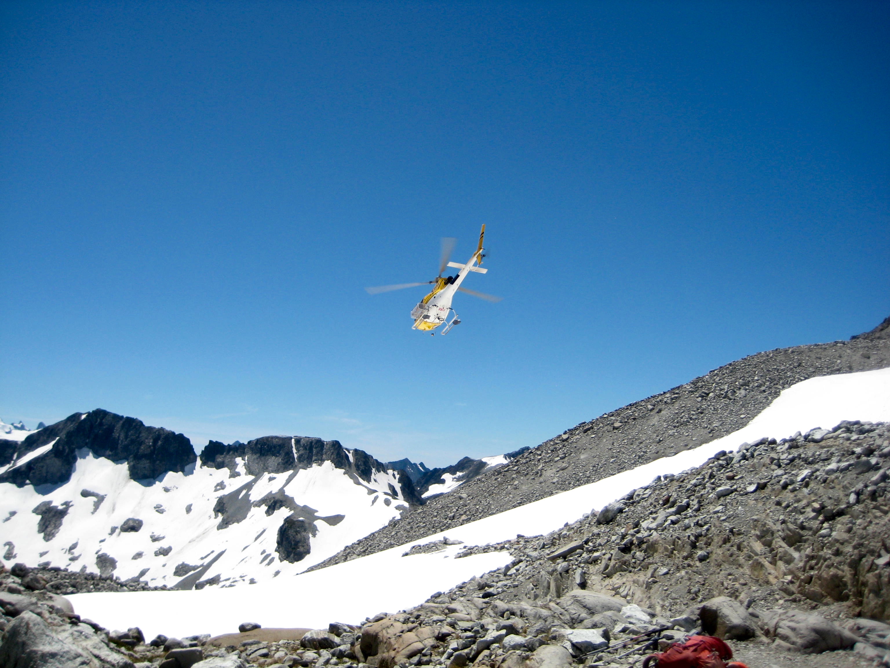 mountain rescue heliocopter flying towrd mountain ridge with scree and snow below in the North Cascades