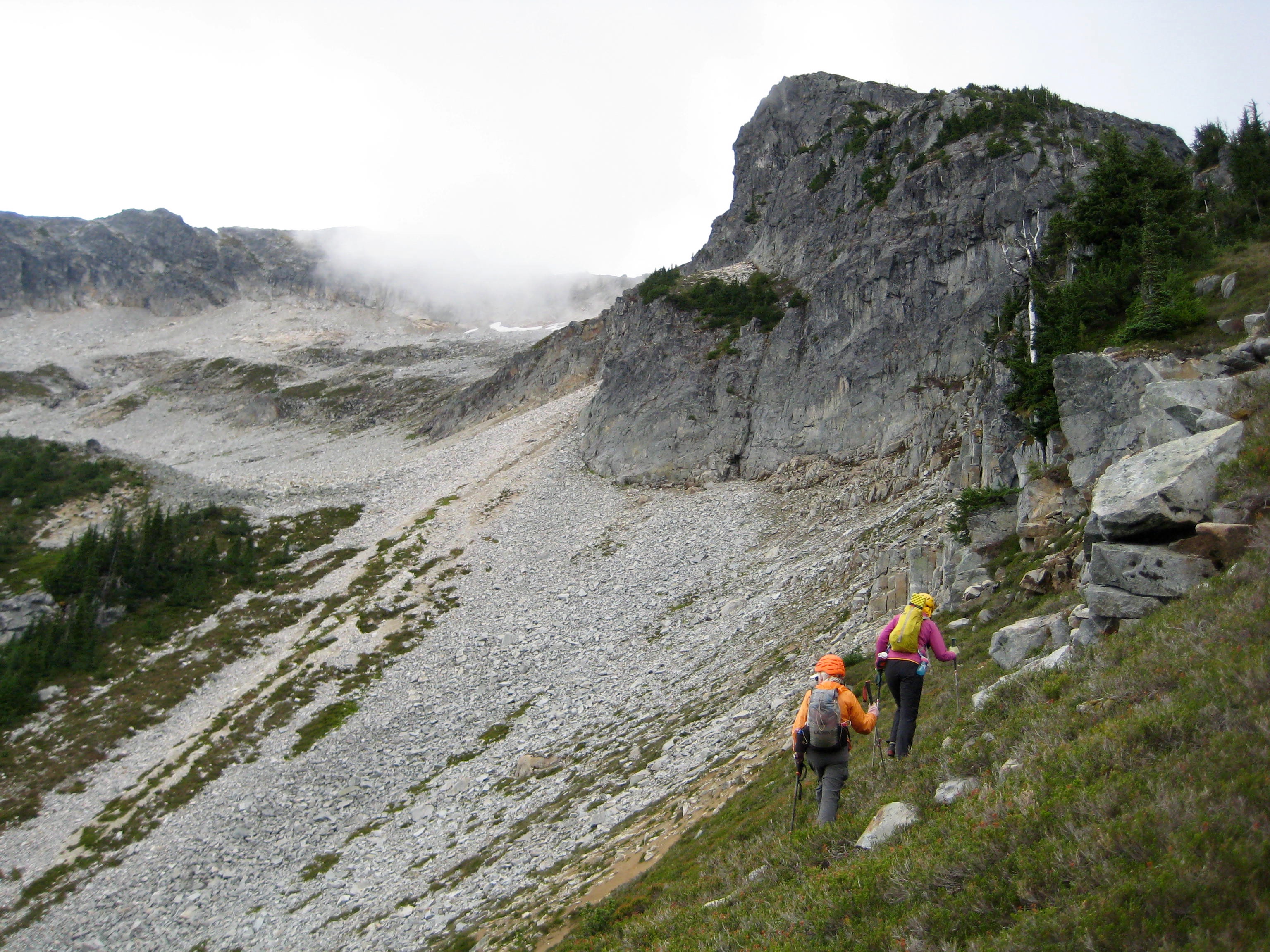 mountain climbers traversing steep grassy slopes heading for scree field under the steep rocky ridge of Bear Mountain in the American Chilliwack Mountains