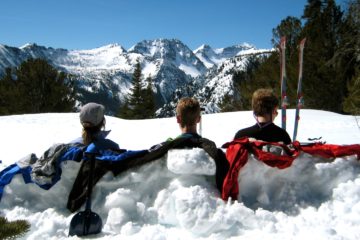 Three skiers sit on sunny McCue Ridge and look at a snowy mountain