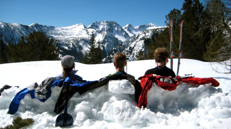 Three skiers sit on sunny McCue Ridge and look at a snowy mountain