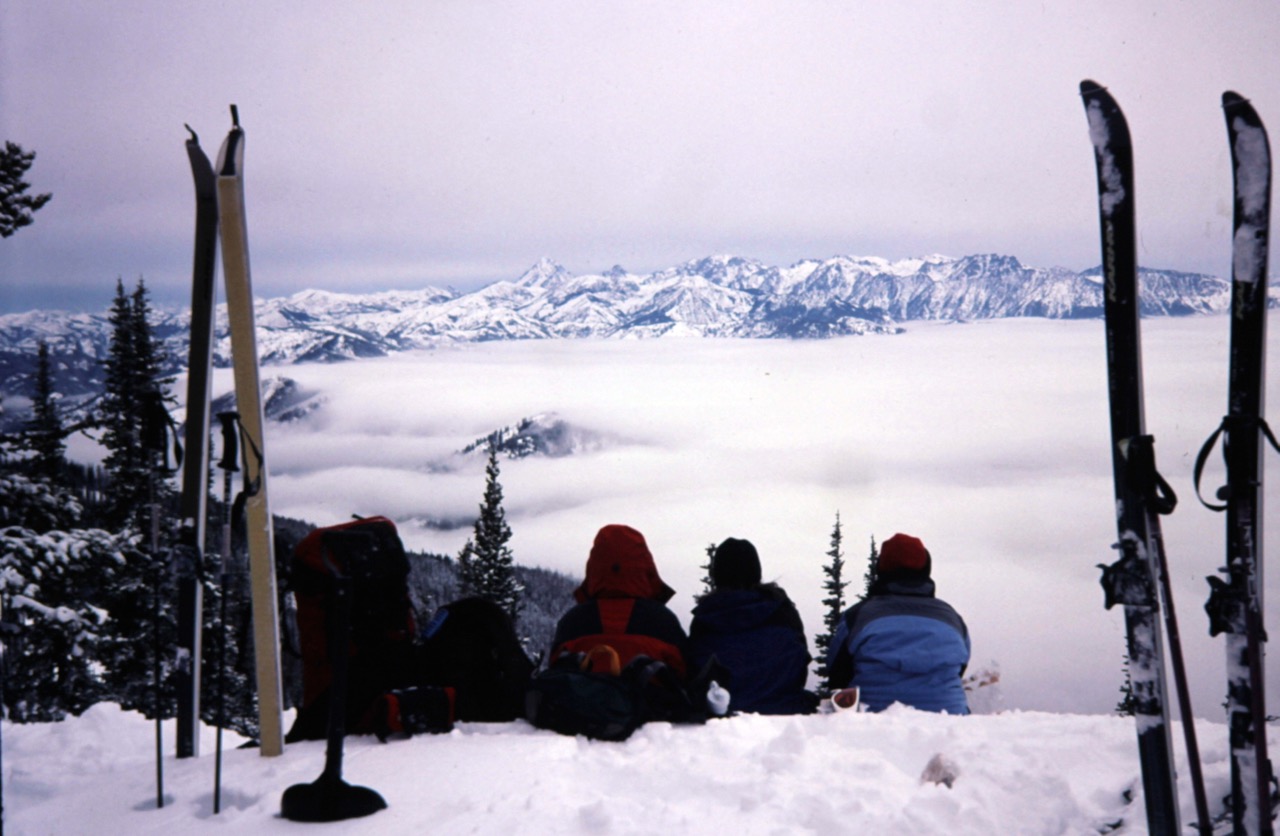 Three skiers sit on Tronsen Head Summit overlooking a foggy valley during lunch break on Tronsen Head Ski Loop