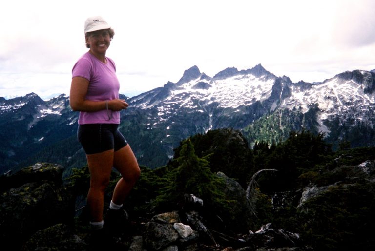 A woman climber stands on Hurricane Peak with a glaciated mountain behind