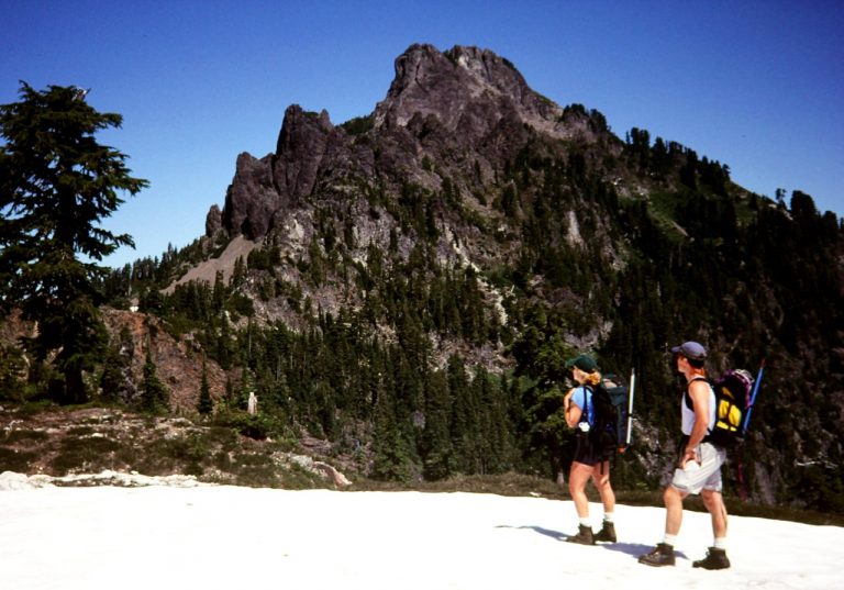 Two hikers stand at a snowy pass and look at a rocky peak