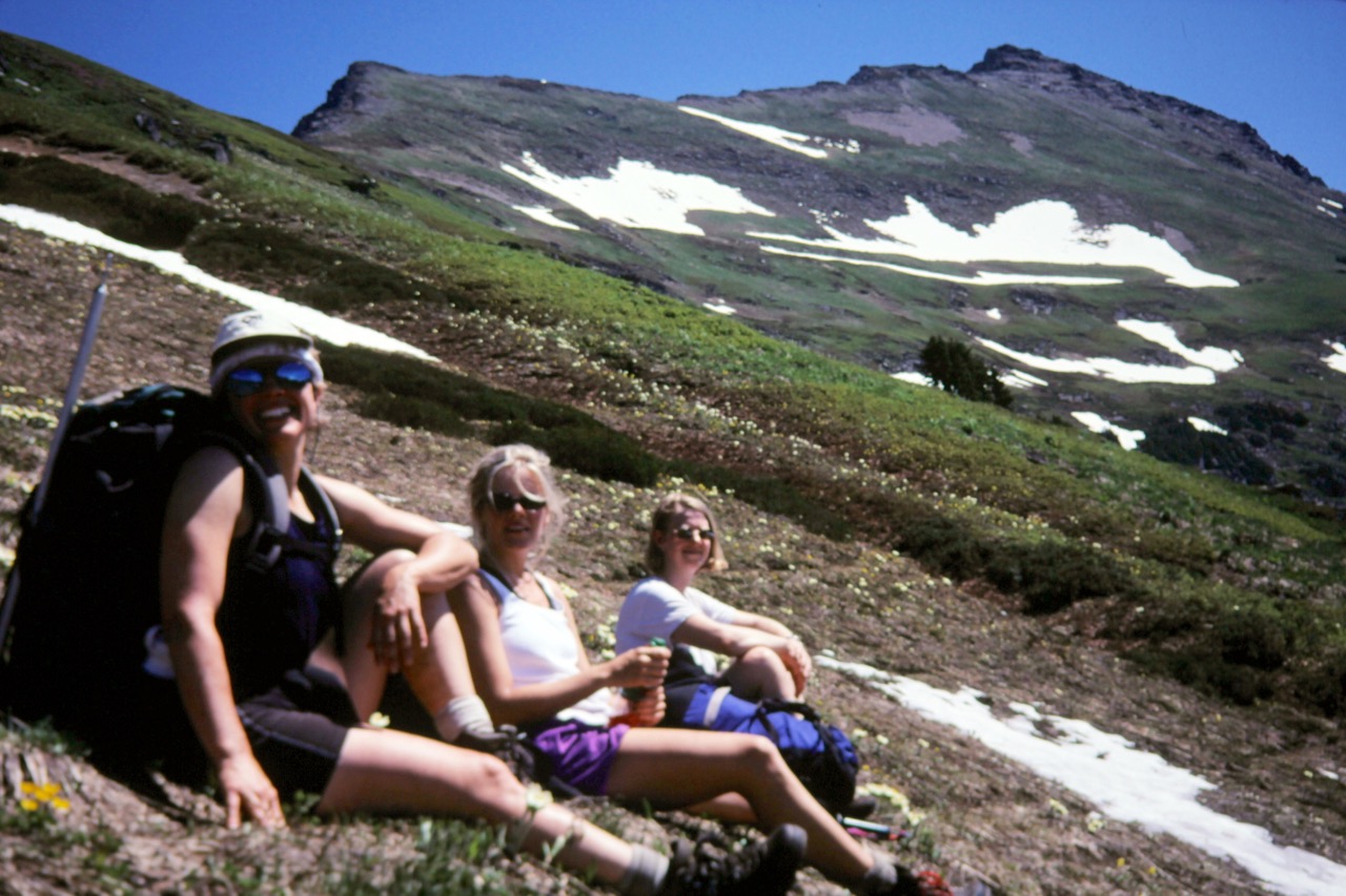 Three hikers sit on a grassy slope below Indian Head Peak during the Little Indian Traverse
