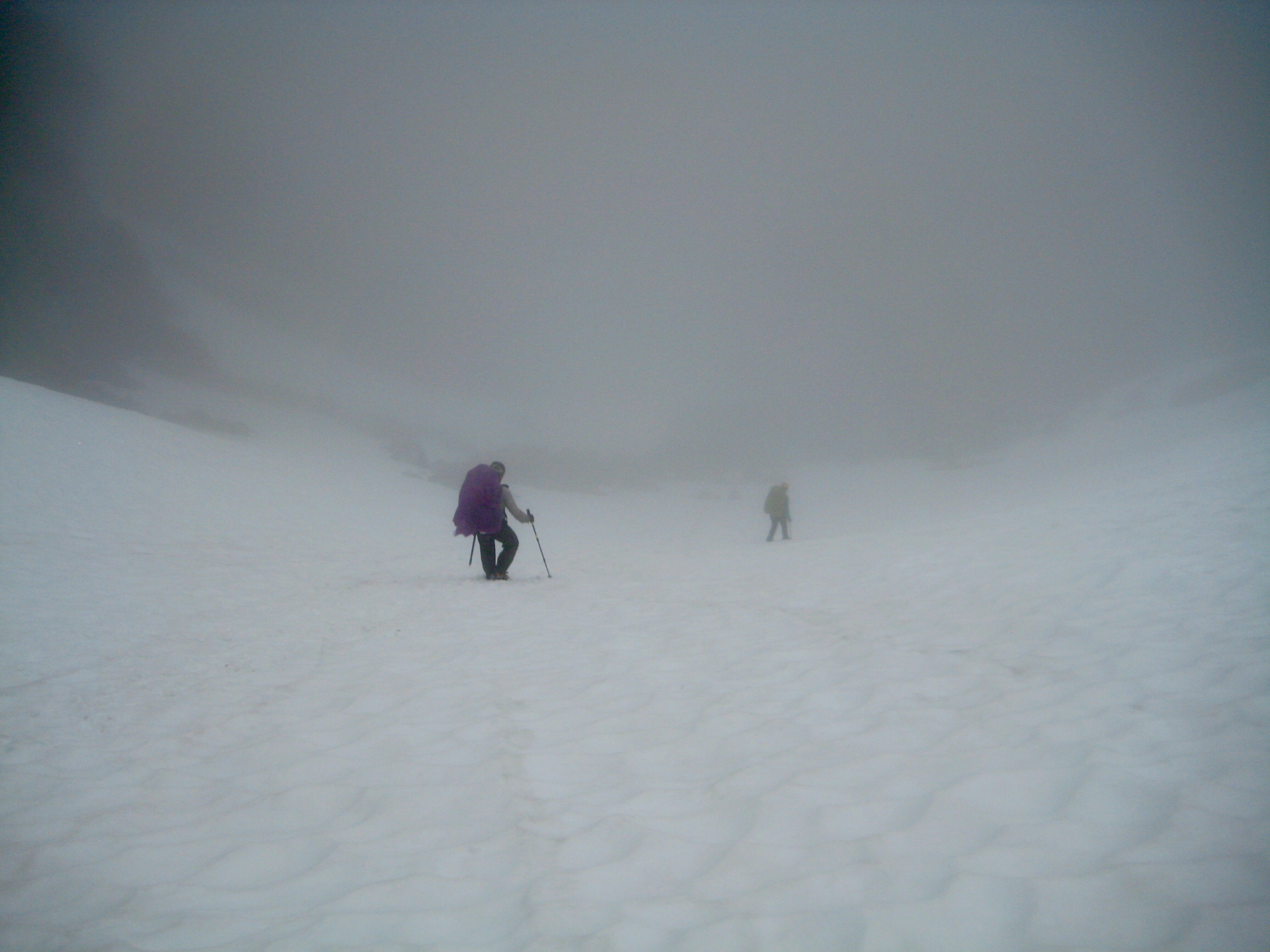 looking down on ountain climber booting down large snow field in Coccyx Col in heavy fog along McAllister Traverse aka Isolation Traverse