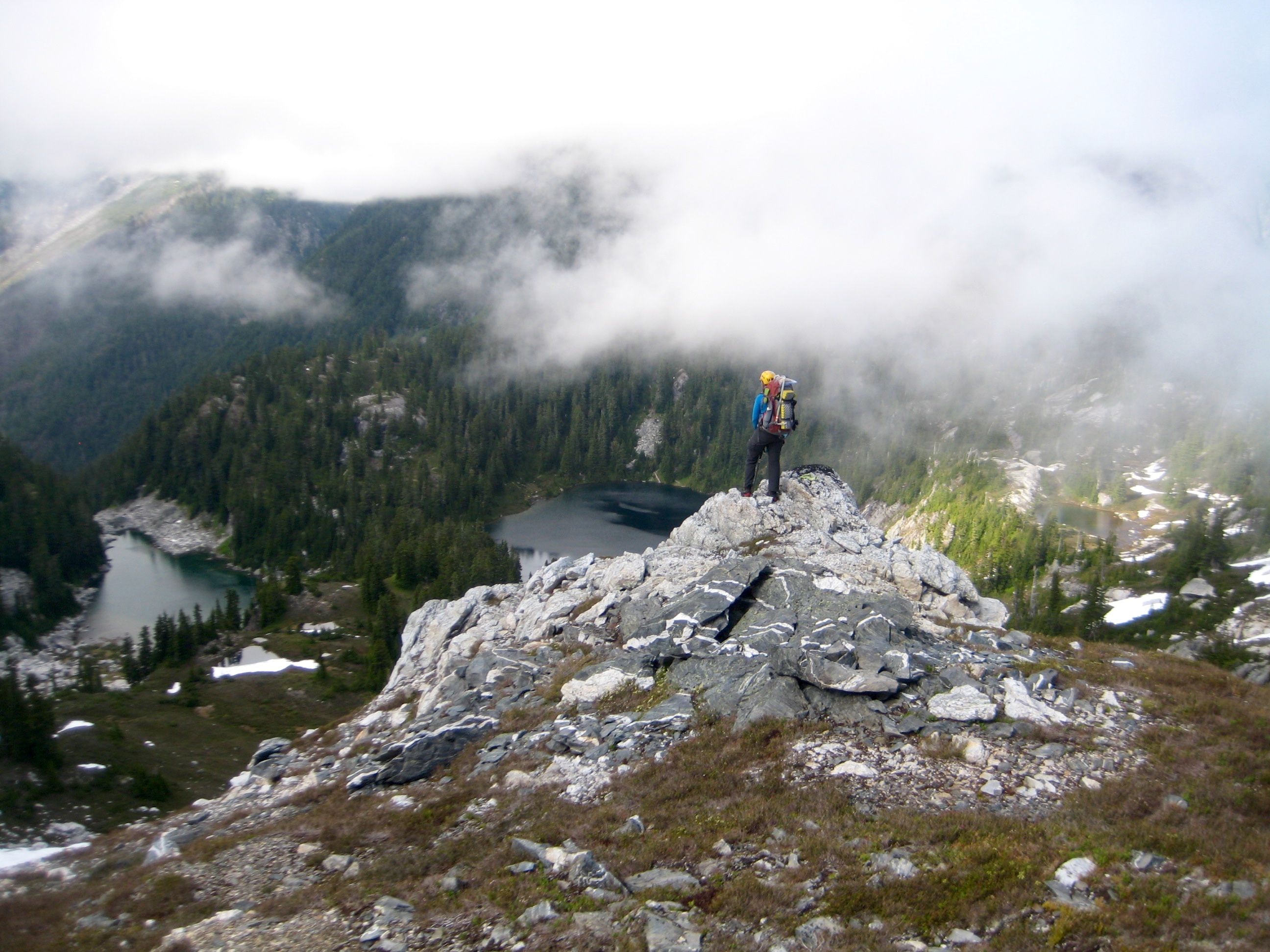 mountain climber standing on rock outcropping on heather ledge looking down on Wilcox Lakes with swirling clouds along the Isolation Traverse