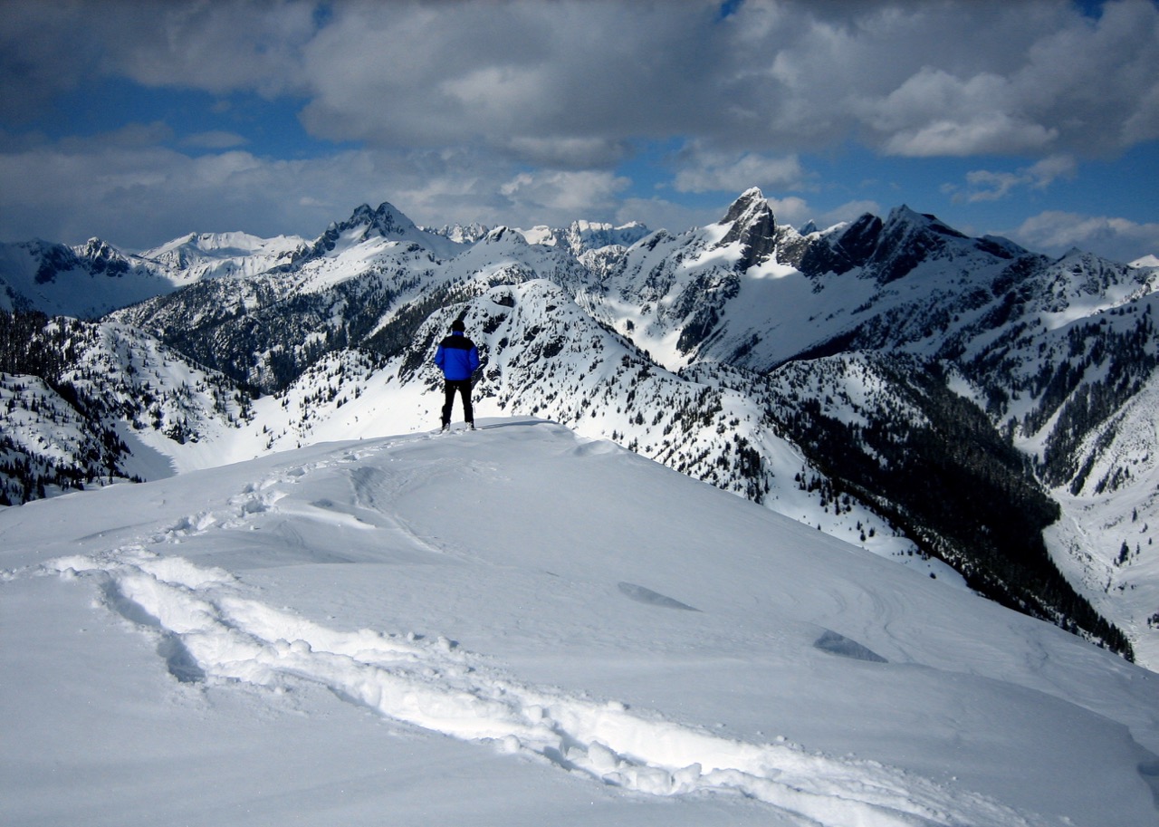 A snowshoe hiker stands on summit of Oakes Peak looking at jagged mountains