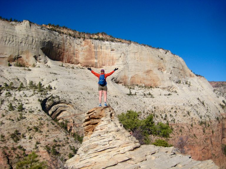 A hiker waves her arms at a sandstone cliff from the summit of Angels Landing in Zion National Park Utah