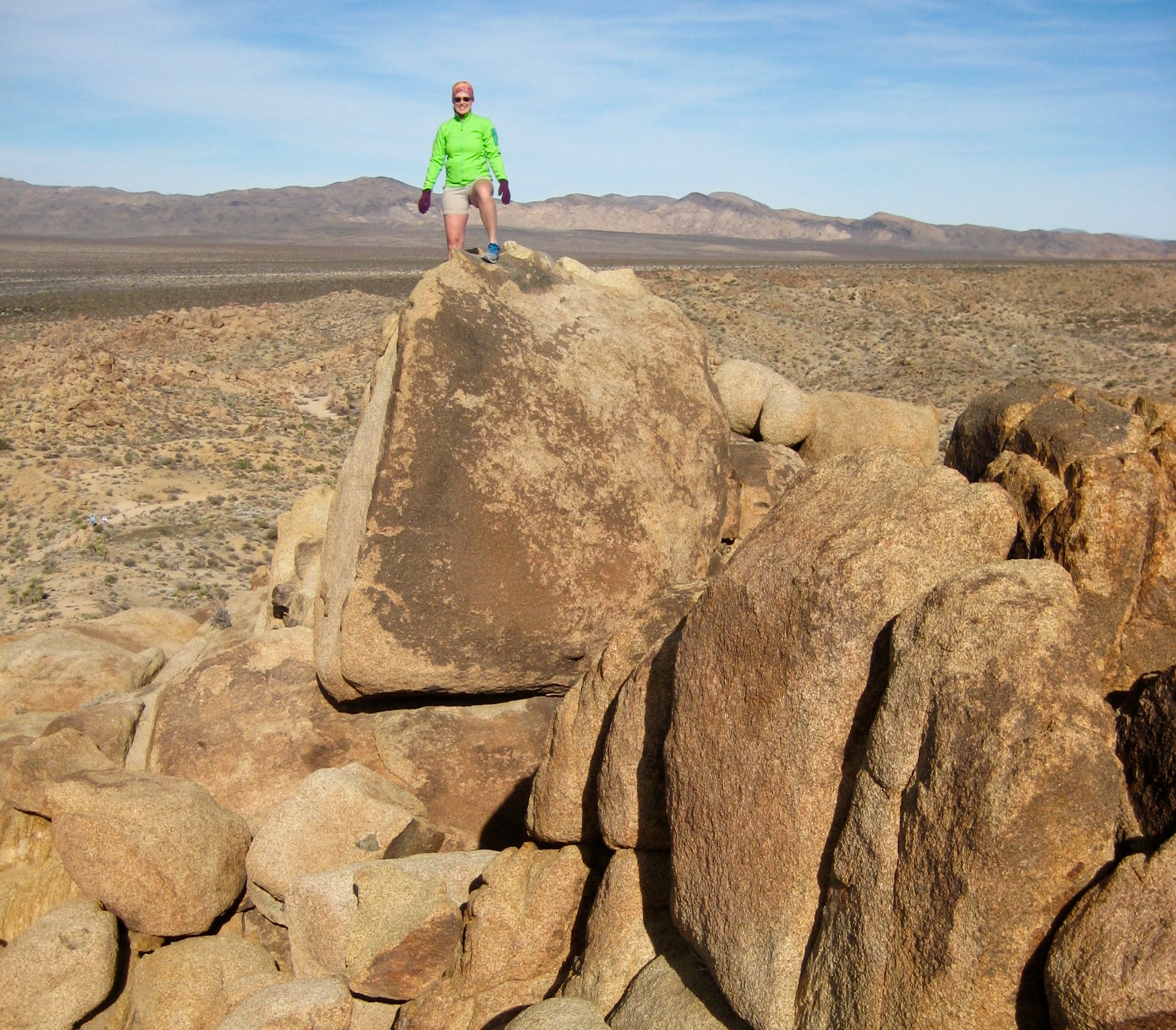hiker stands on top of the large, blouder summit of Mastodon Peak with Joshua Tree National Park in the background 