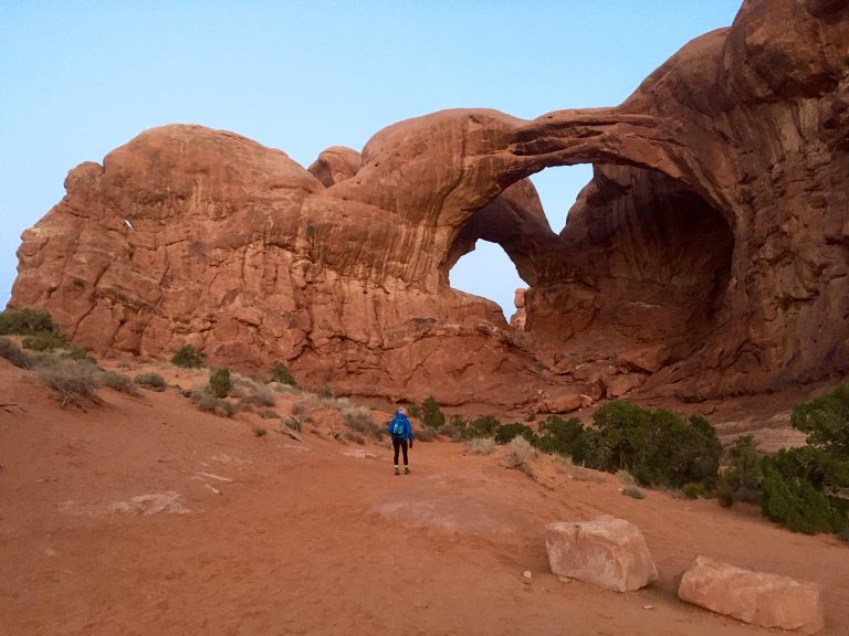 A hiker walks toward Double Arch in Arches National Park before sunrise